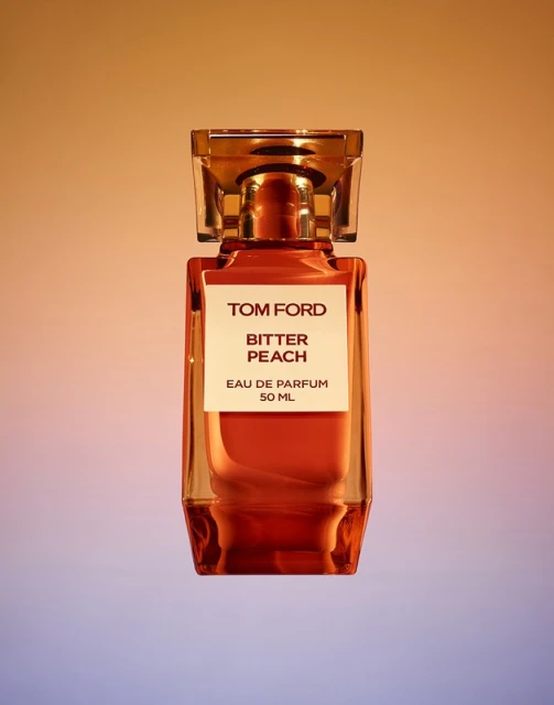 Tom Ford Bitter Peach perfume bottle. The transparent glass bottle has a amber liquid inside and a clear geometric stopper.