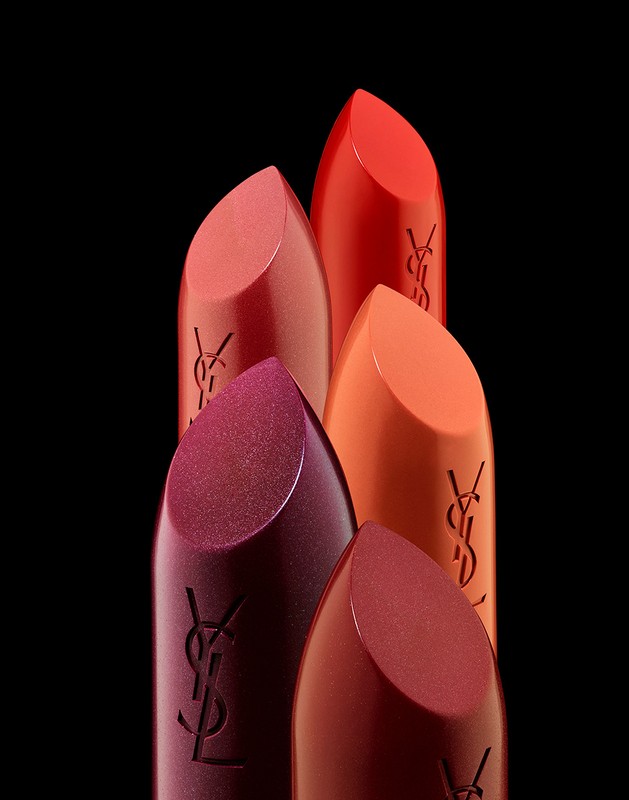 A group of Yves Saint Laurent lipsticks in various shades of red, pink, and plum, arranged against a stark black backdrop.