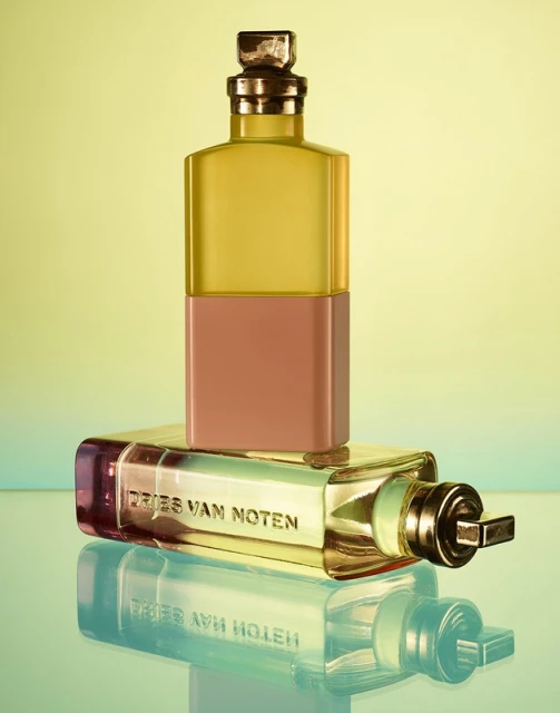 Two Dries Van Noten perfume bottles are stacked against a graduated yellow-green background, showcasing their unique shapes and design.