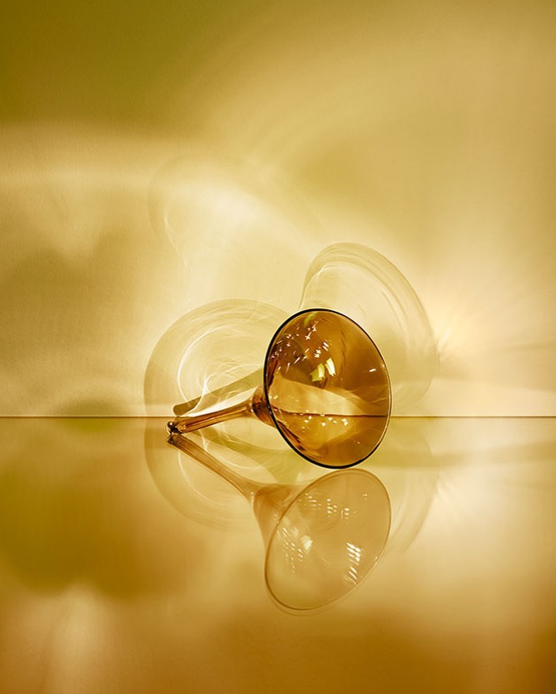 Still life of an amber-colored glass trumpet vase with its reflection against a tan-yellow background, illuminated by studio lighting.