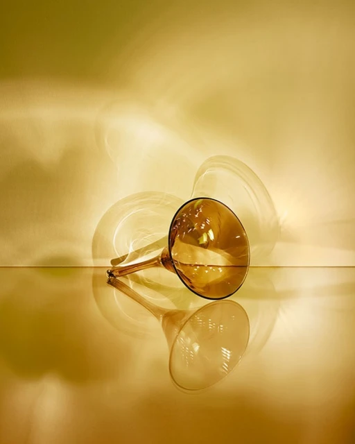 Still life of an amber-colored glass trumpet vase with its reflection against a tan-yellow background, illuminated by studio lighting.