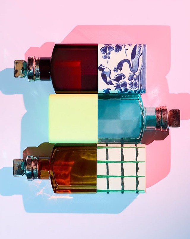 Three colorful perfume bottles with patterned caps are arranged horizontally against a gradient pink and blue background in a studio shot.