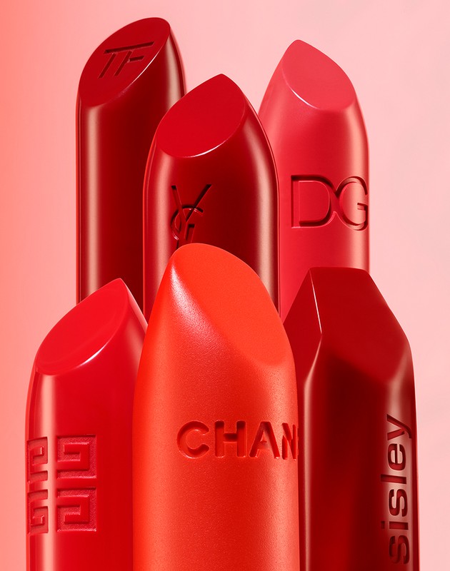 Close-up studio shot of six red lipsticks from various luxury cosmetic brands, including Chanel, Dior, and Tom Ford, on a pale pink background.