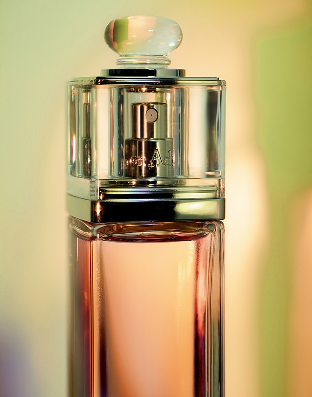 Close-up studio shot of a transparent perfume bottle with a silver cap, filled with peach-colored liquid, against a blurred background.