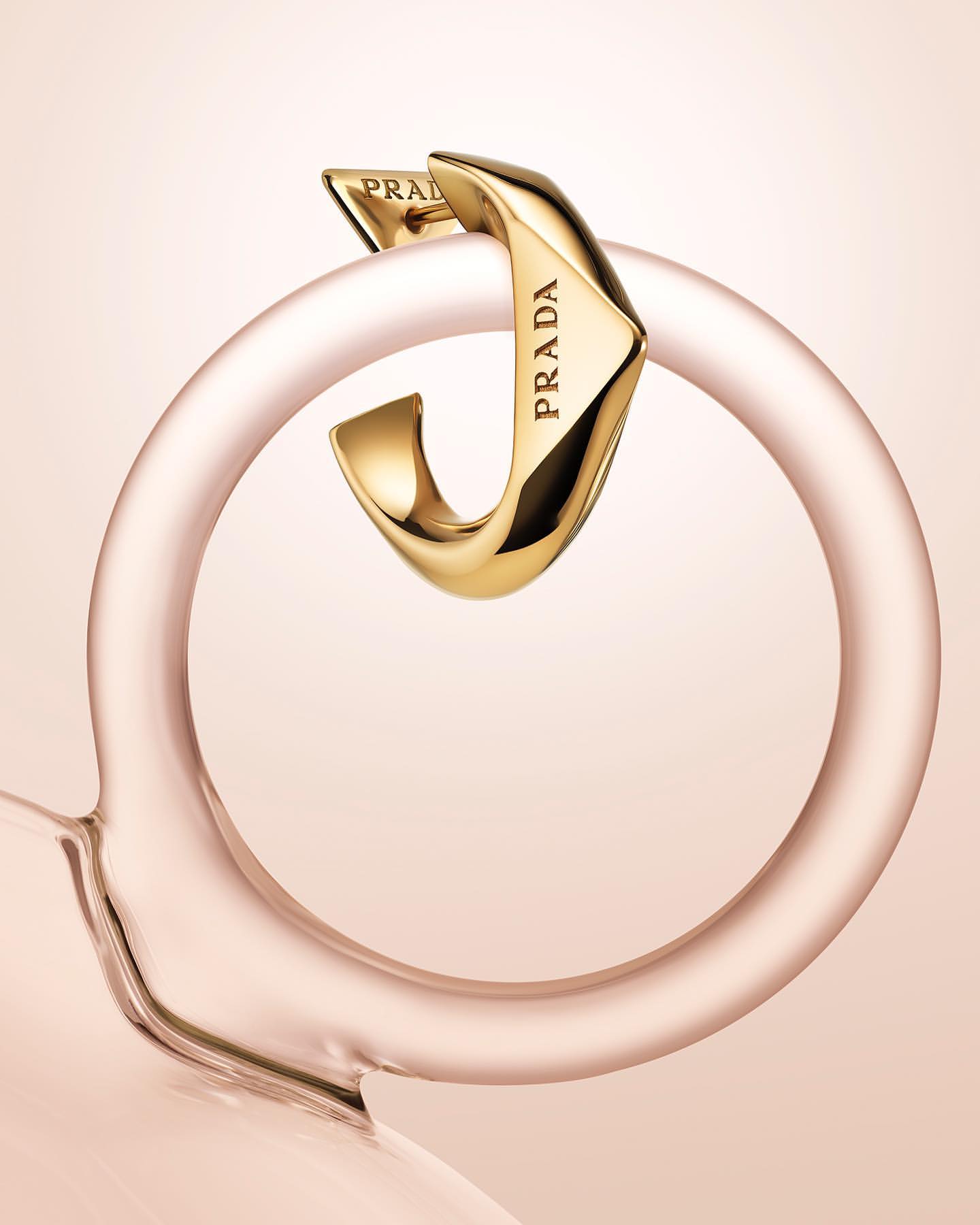 Close-up studio shot of a gold-toned Prada earring looped around a transparent pink glass ring. Light pink background.