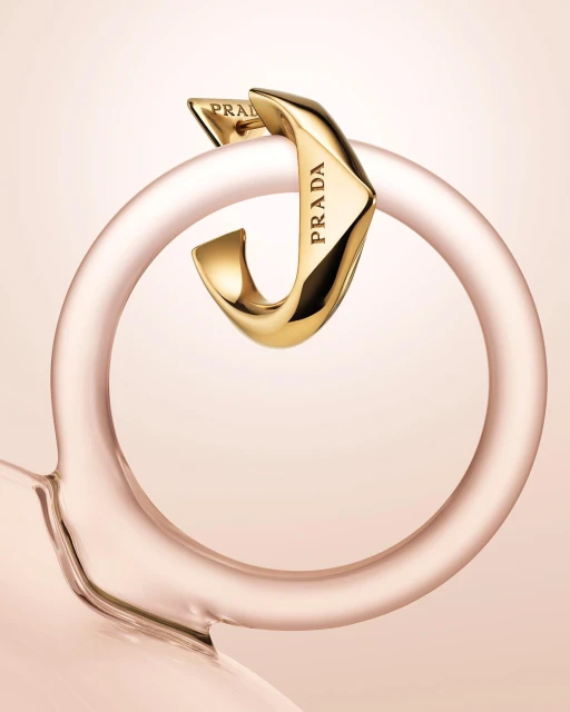 Close-up studio shot of a gold-toned Prada earring looped around a transparent pink glass ring. Light pink background.