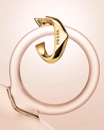 Close-up studio shot of a gold-toned Prada earring looped around a transparent pink glass ring. Light pink background.