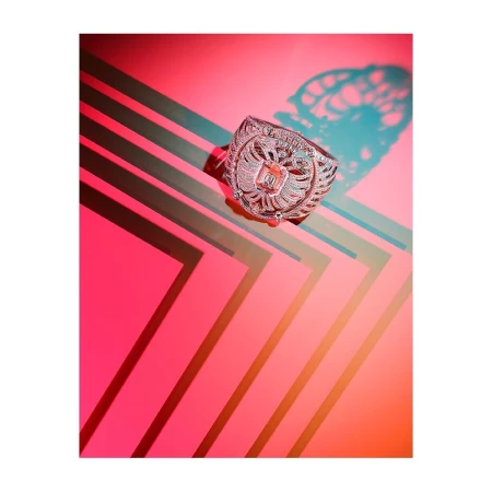 A silver ring featuring an eagle design is displayed on a vibrant pink background with geometric lines and contrasting shadows.