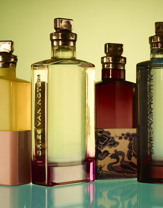 Four Dries Van Noten perfume bottles stand side-by-side, showcasing different colors and designs against a soft yellow-green background, with light reflections.