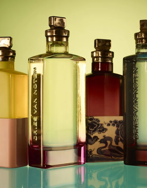 Four Dries Van Noten perfume bottles stand side-by-side, showcasing different colors and designs against a soft yellow-green background, with light reflections.