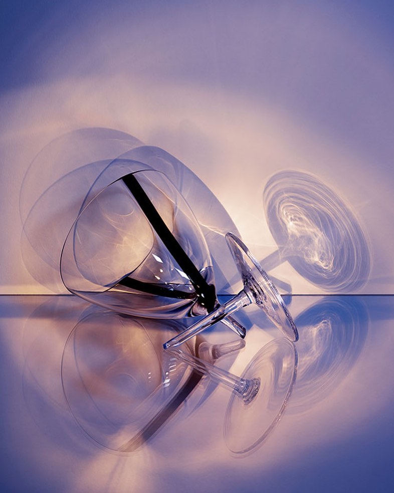Abstract studio photograph of an empty clear wine glass lying on its side on a reflective surface with a gradient purple background and long shadows.