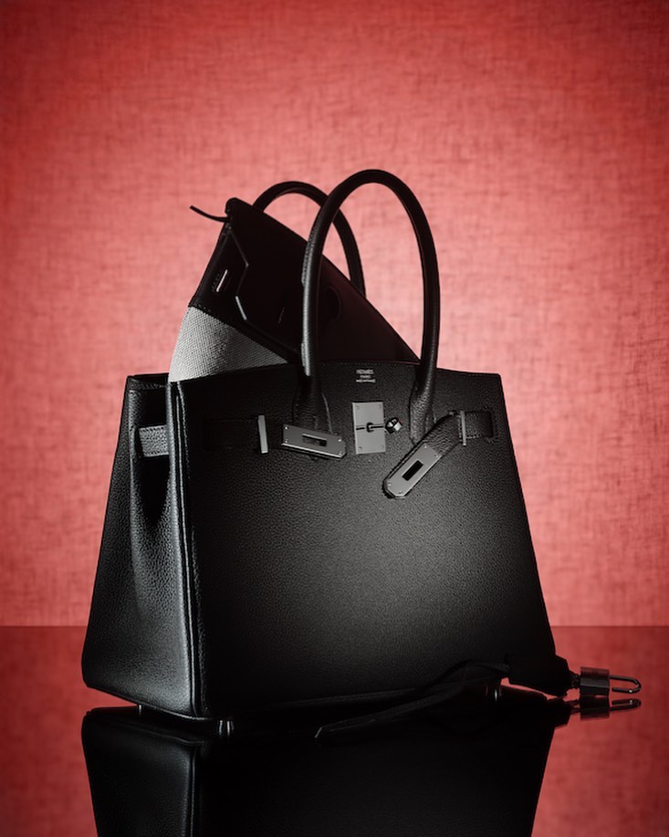 Still life shot of two black leather handbags with silver hardware, set against a textured red background with a reflective surface.