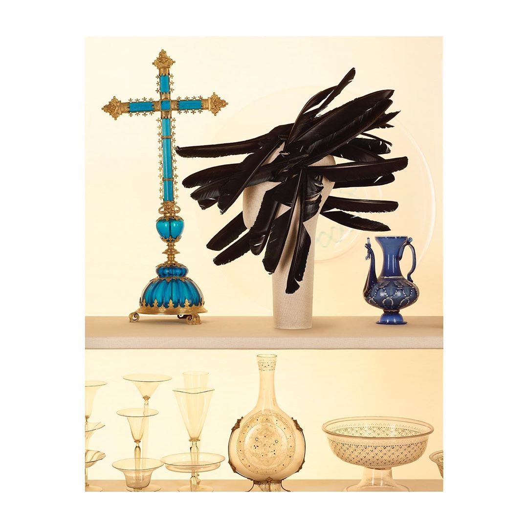 Still life of ornamental cross, feather arrangement, and decorative vases displayed on tiered shelving against a pale yellow backdrop.