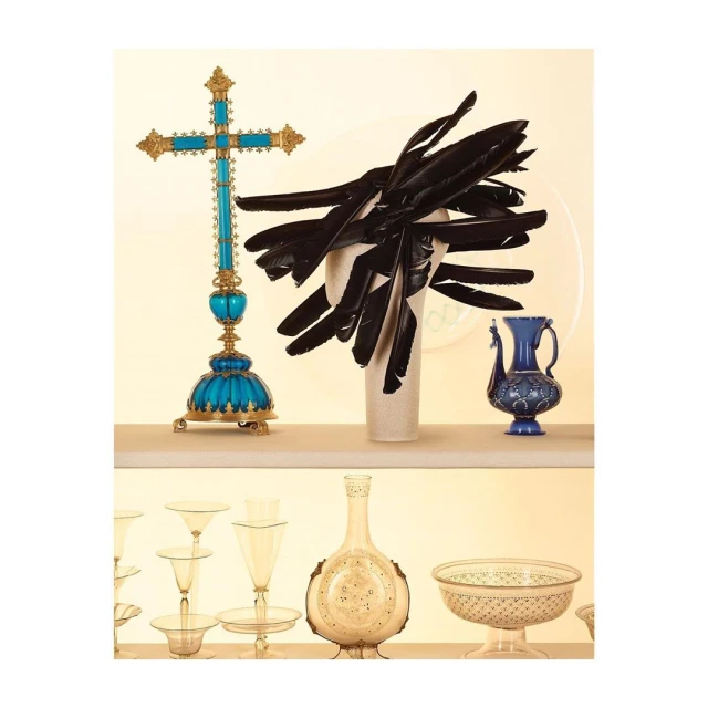 Still life of ornamental cross, feather arrangement, and decorative vases displayed on tiered shelving against a pale yellow backdrop.