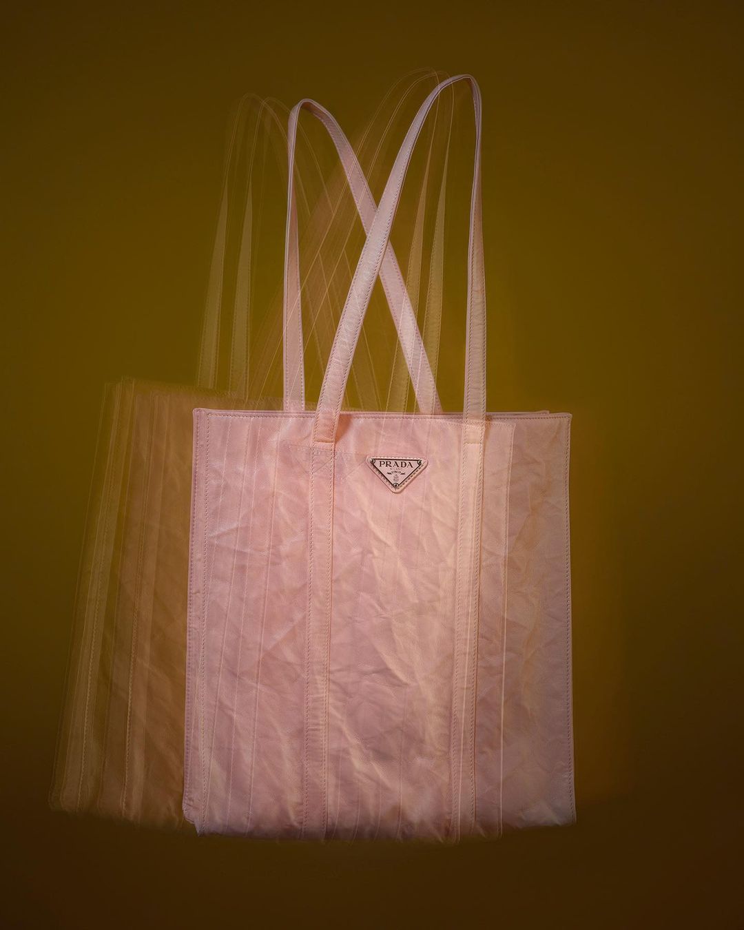 Prada blush-toned tote bag featuring long, light-colored shoulder straps and a triangular logo, captured with motion blur against an olive background.