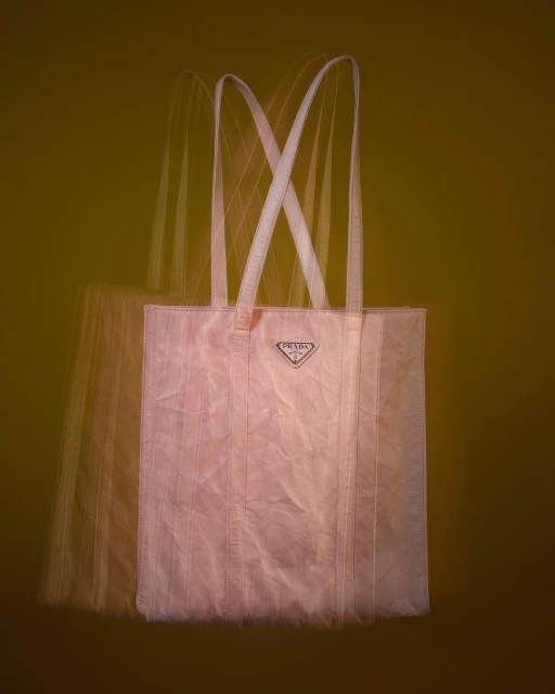 Prada blush-toned tote bag featuring long, light-colored shoulder straps and a triangular logo, captured with motion blur against an olive background.