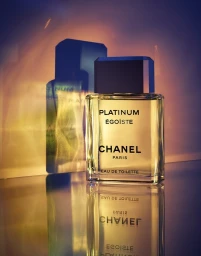 Chanel Platinum Egoiste perfume bottle standing on a reflective surface with warm lighting and a blue shadow projection behind it.