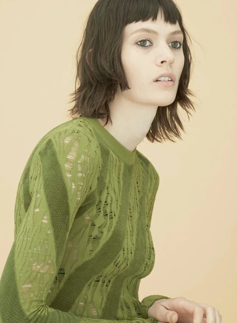 Close-up of a stylish young woman with dark, wavy hair and a green sweater, posing against a soft beige background in a studio.