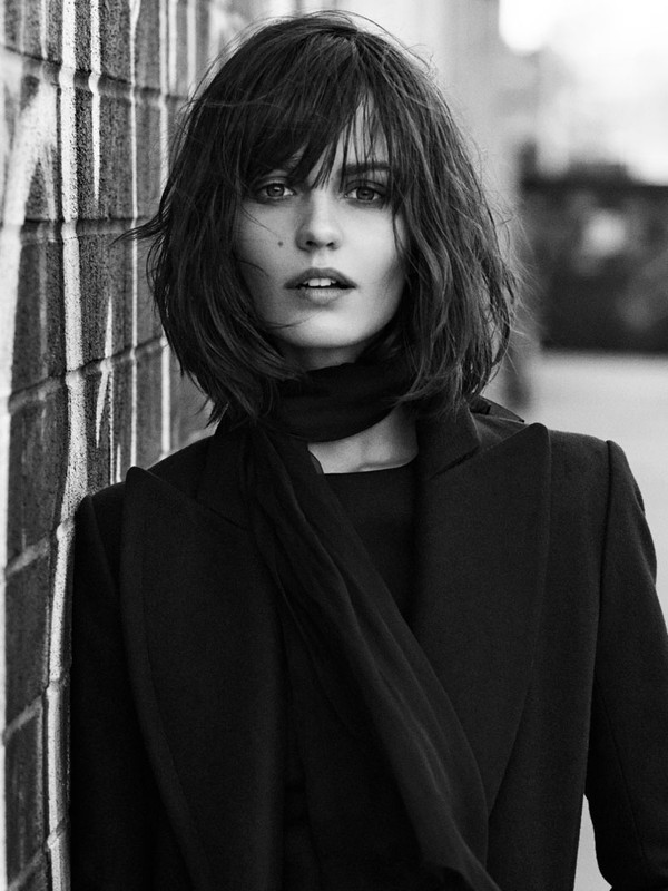 Monochrome portrait of a stylish woman with bob haircut in a black coat and scarf, leaning against a graffiti-covered brick wall.