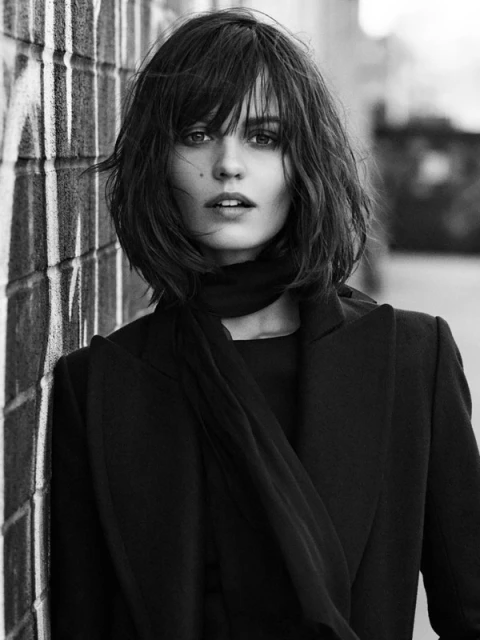 Monochrome portrait of a stylish woman with bob haircut in a black coat and scarf, leaning against a graffiti-covered brick wall.