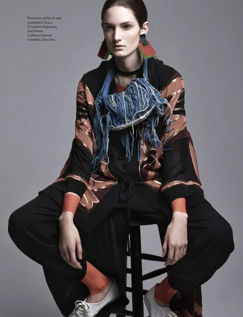 A woman in fashion attire sits on a black stool, wearing a patterned kimono, wide-legged pants, statement earrings, and chunky necklace.