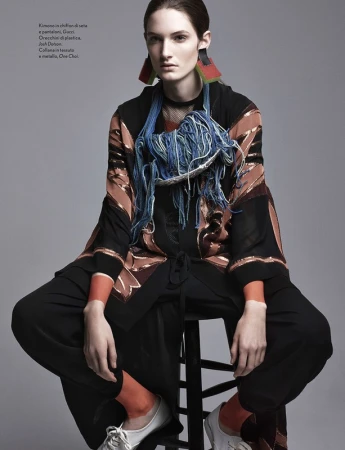 A woman in fashion attire sits on a black stool, wearing a patterned kimono, wide-legged pants, statement earrings, and chunky necklace.