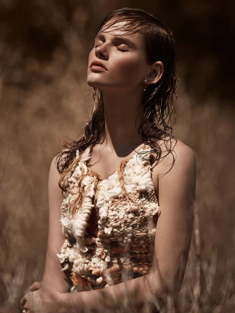 Fashion shot of a fair-skinned adult woman with wet hair, eyes closed, wearing a cream-colored woven top, photographed outdoors in natural light.