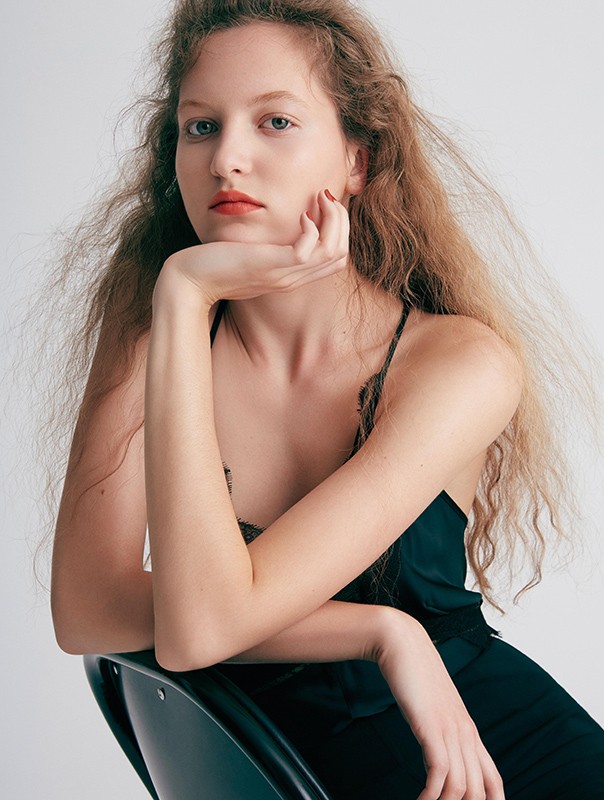 Close-up portrait of a woman with long wavy blonde hair resting her chin on her hand, wearing a dark green top against a white wall.