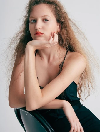 Close-up portrait of a woman with long wavy blonde hair resting her chin on her hand, wearing a dark green top against a white wall.
