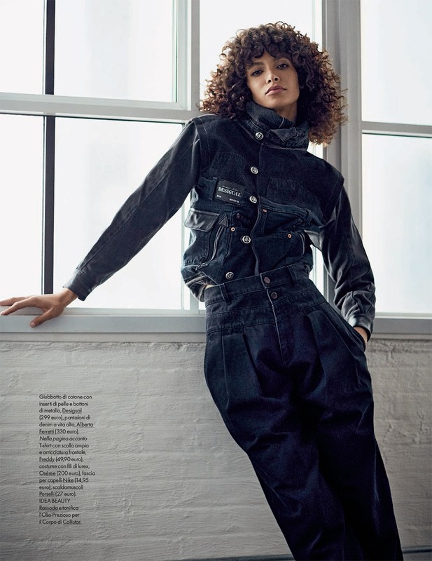 Model with curly hair leaning against a large window wearing a dark denim jacket and high-waisted trousers.