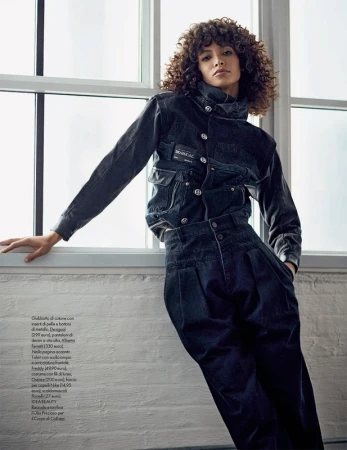 Model with curly hair leaning against a large window wearing a dark denim jacket and high-waisted trousers.