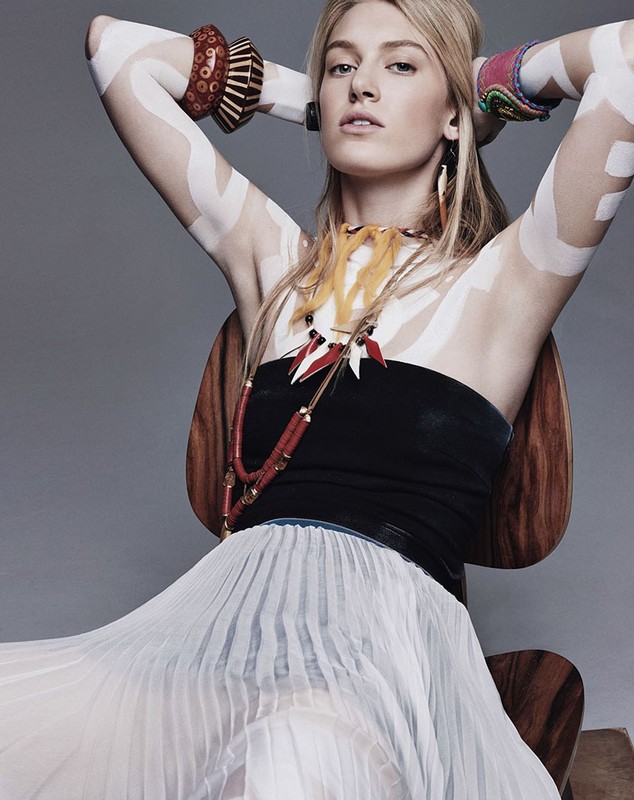 Blonde woman with geometric body paint, tribal jewelry, and a flowing skirt sits in a wooden chair in a studio setting.