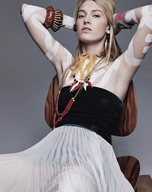 Blonde woman with geometric body paint, tribal jewelry, and a flowing skirt sits in a wooden chair in a studio setting.