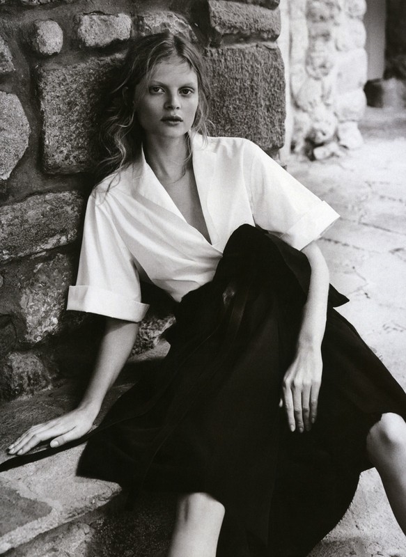 A fair-skinned blonde woman sits on stone steps in black and white fashion portrait with white top and black skirt.