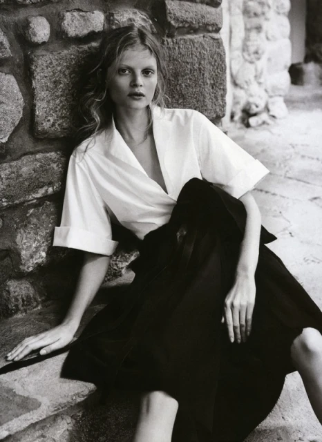 A fair-skinned blonde woman sits on stone steps in black and white fashion portrait with white top and black skirt.