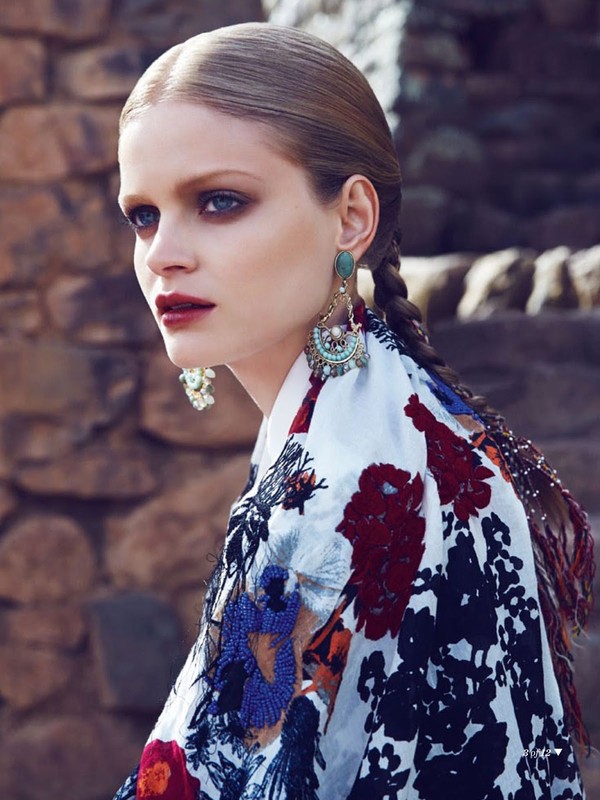 Portrait of an adult female model with fair skin wearing a floral-print jacket and elaborate earrings against a stone wall backdrop.
