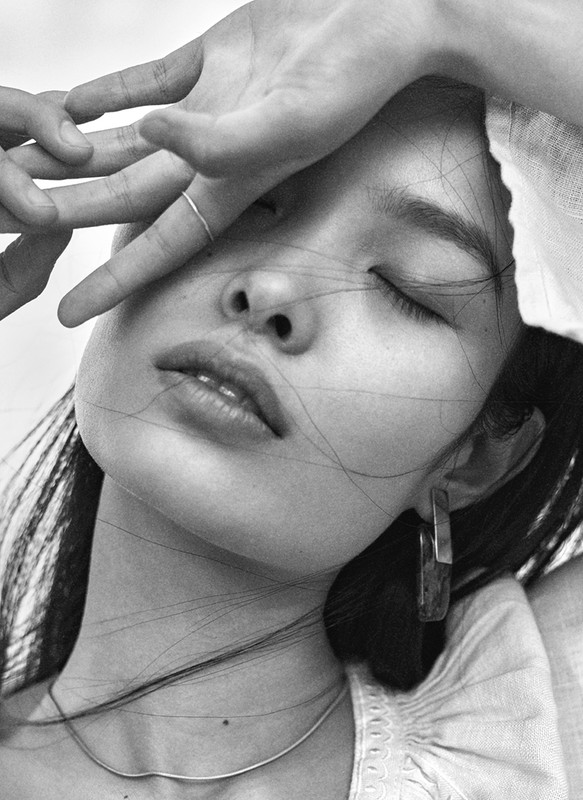 Monochrome portrait of a young Asian woman with eyes closed, hand near face, accessorized with a ring, necklace, and earrings.