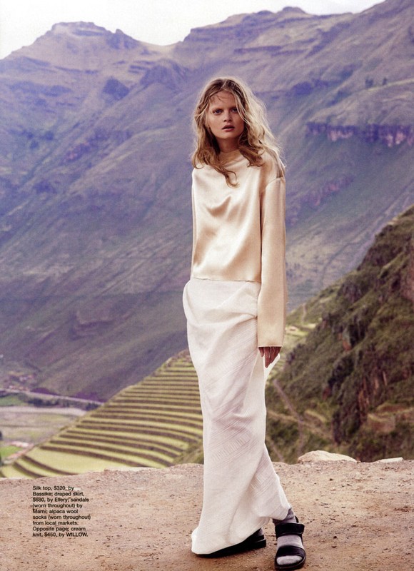 A woman in a long white skirt and gold blouse poses in front of a mountain landscape with terraced fields on the slopes.