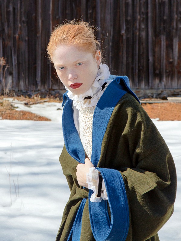 Redhead woman in dark green and blue coat stands outside in snowy area with rustic building in background, fashion editorial.