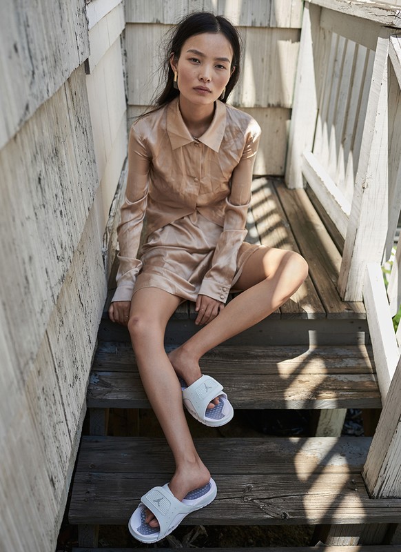 Asian woman in silk dress and slide sandals sits on wooden steps outdoors with neutral expression in natural light in a fashion pose.