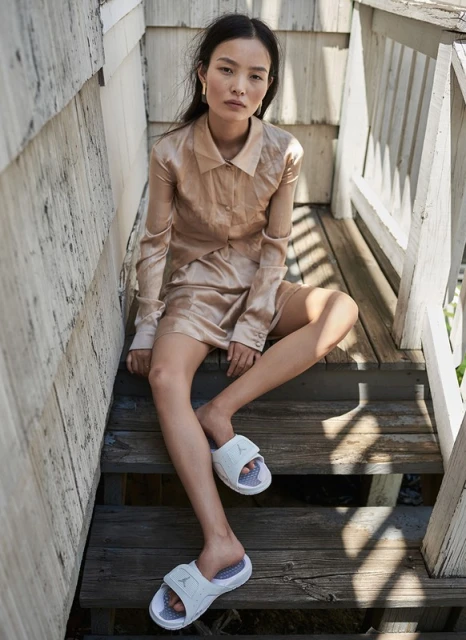 Asian woman in silk dress and slide sandals sits on wooden steps outdoors with neutral expression in natural light in a fashion pose.