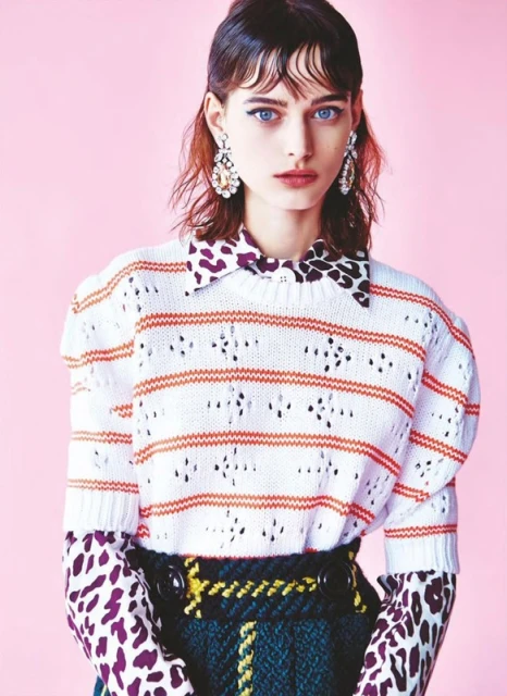 Fashion portrait of a female model with light skin, wearing patterned top and skirt, accessorized with earrings, against a pink background.