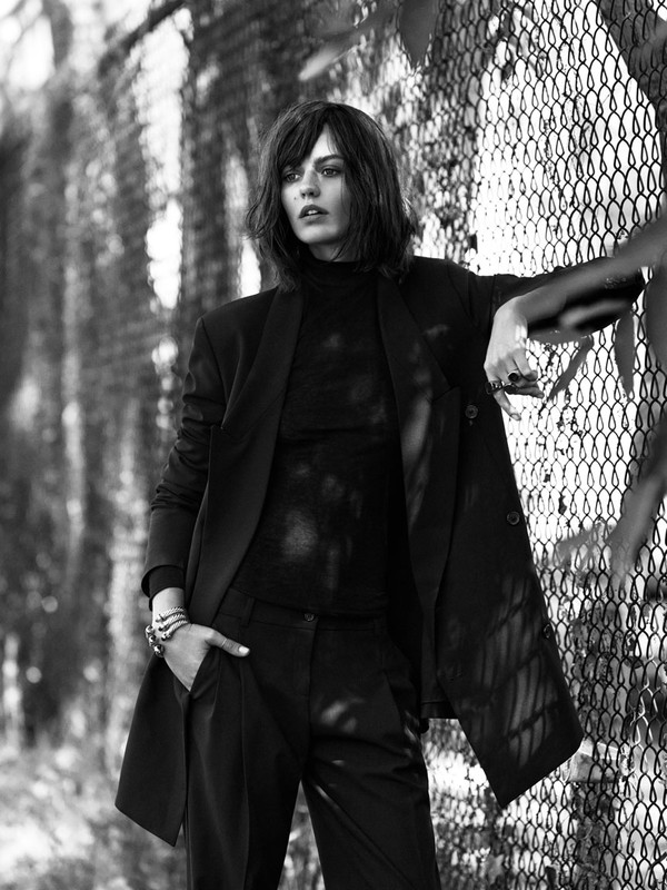 A stylish brunette woman in a black suit leans against a chain-link fence in a black and white outdoor fashion shot.