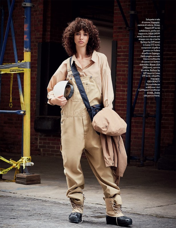 Fashion editorial shot of a woman in beige coveralls holding a hard hat in an outdoor urban environment with scaffolding.