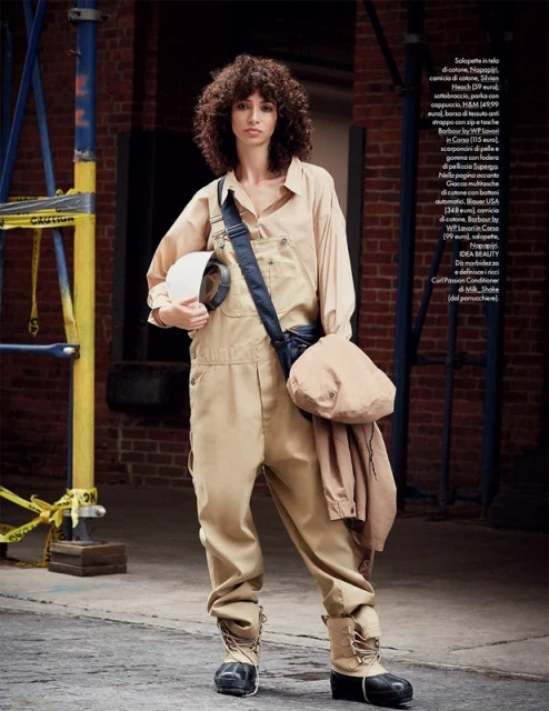 Fashion editorial shot of a woman in beige coveralls holding a hard hat in an outdoor urban environment with scaffolding.