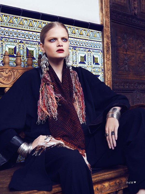 Fashion shot of a woman with fair skin and blue eyes wearing a black outfit and patterned scarf, sitting against a decorative tile wall.