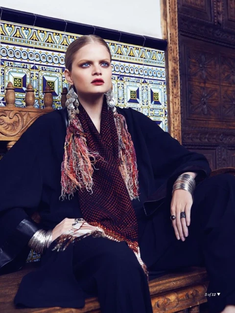 Fashion shot of a woman with fair skin and blue eyes wearing a black outfit and patterned scarf, sitting against a decorative tile wall.