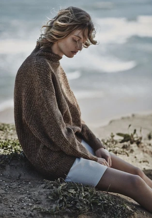 Woman in a brown turtleneck sweater seated outdoors near a coastline with choppy water in the background