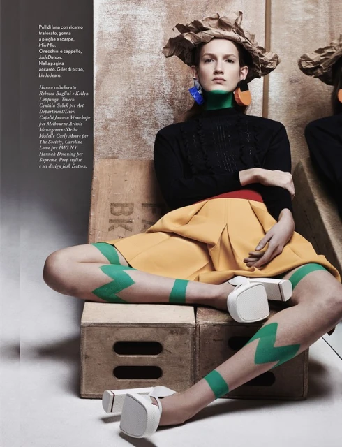 Fashion model in a paper hat, cable-knit sweater, mustard skirt, and white shoes, sitting on wooden crates in a studio environment.