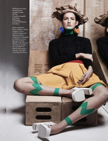 Fashion model in a paper hat, cable-knit sweater, mustard skirt, and white shoes, sitting on wooden crates in a studio environment.